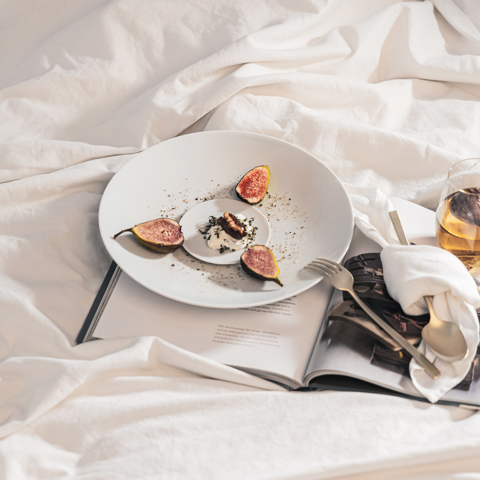 TAC plate and bread plate garnished with figs and dessert lie on a table sheet along with an open book, cutlery, white napkin and glass.